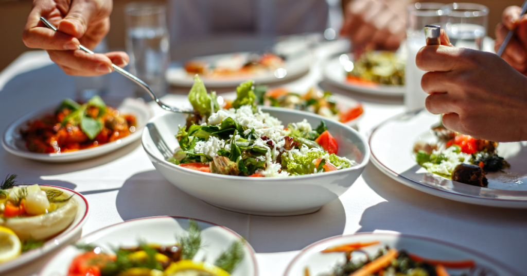 Close-up of people sharing a meal with fresh salads, vegetables, and healthy dishes, emphasizing mindful eating and intuitive eating practices.