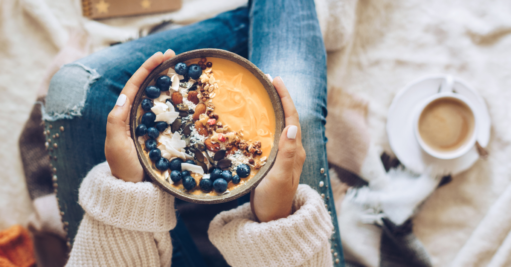 A woman holding a smoothie bowl topped with fresh blueberries, coconut flakes, and granola, sitting in a cozy setting with a cup of coffee.